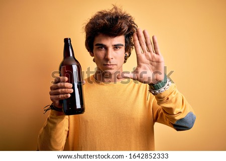 Young handsome man drinking bottle of beer standing over isolated yellow background with open hand doing stop sign with serious and confident expression, defense gesture