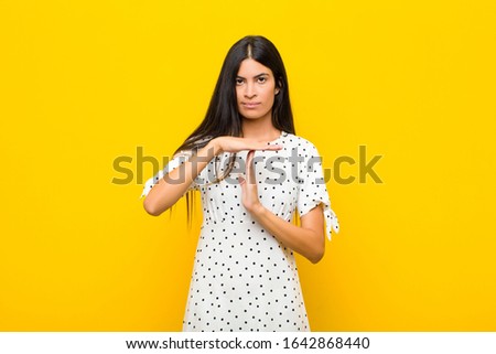 young pretty latin woman looking serious, stern, angry and displeased, making time out sign against flat wall