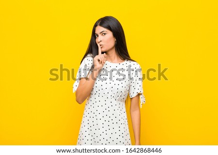 young pretty latin woman asking for silence and quiet, gesturing with finger in front of mouth, saying shh or keeping a secret against flat wall