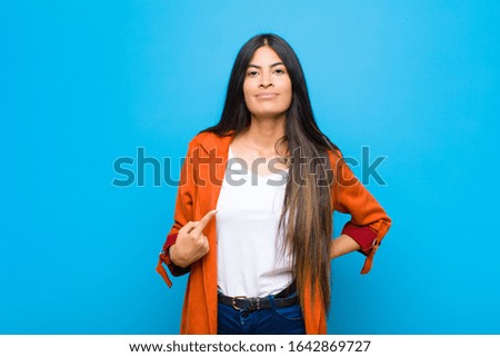 young pretty latin woman looking proud, confident and happy, smiling and pointing to self or making number one sign against flat wall