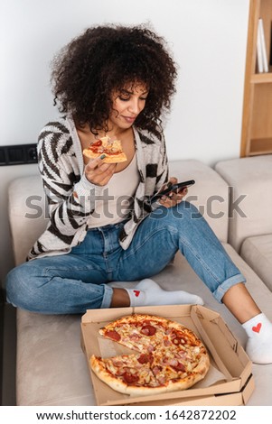 Image of optimistic positive pretty young african woman indoors at home eat pizza chatting by phone.