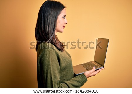Young brunette woman with blue eyes working using computer laptop over yellow background looking to side, relax profile pose with natural face and confident smile.