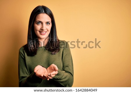 Young brunette woman with blue eyes wearing green casual sweater over yellow background Smiling with hands palms together receiving or giving gesture. Hold and protection