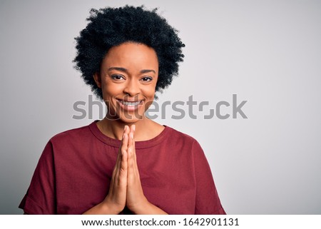 Young beautiful African American afro woman with curly hair wearing casual t-shirt standing praying with hands together asking for forgiveness smiling confident.
