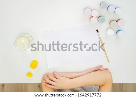 Top view on a white table, a sheet of paper, paints, brush, hands of a child. A child waiting for the start of drawing. Space for text or picture.
