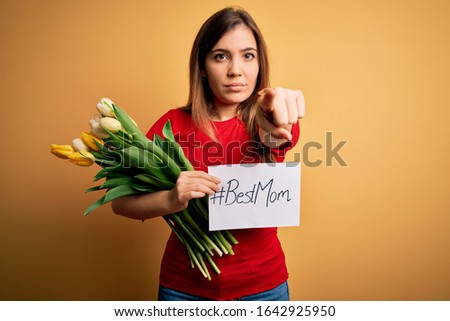 Beautiful woman holding paper with best mom message and tulips celebrating mothers day pointing with finger to the camera and to you, hand sign, positive and confident gesture from the front