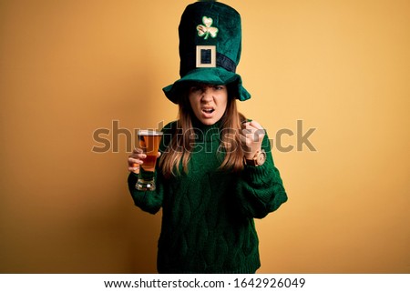 Young beautiful woman wearing green hat drinking glass of beer on saint patricks day angry and mad raising fist frustrated and furious while shouting with anger. Rage and aggressive concept.