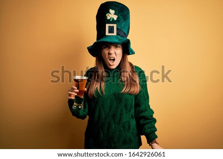 Young beautiful woman wearing green hat drinking glass of beer on saint patricks day angry and mad screaming frustrated and furious, shouting with anger. Rage and aggressive concept.
