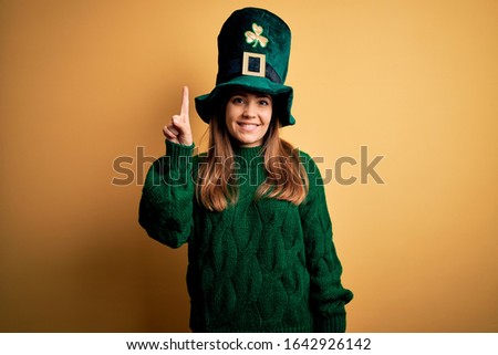 Young beautiful brunette woman wearing green hat on st patricks day celebration showing and pointing up with finger number one while smiling confident and happy.