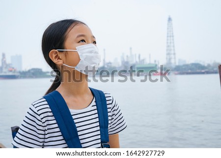 Virus protection concept - asian little girl wearing protective mask, standing near river in smog day. Pollution every where. Looking at the sky.