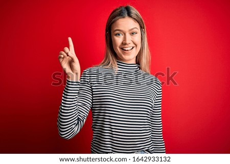 Young beautiful blonde woman wearing casual striped sweater over red isolated background showing and pointing up with finger number one while smiling confident and happy.
