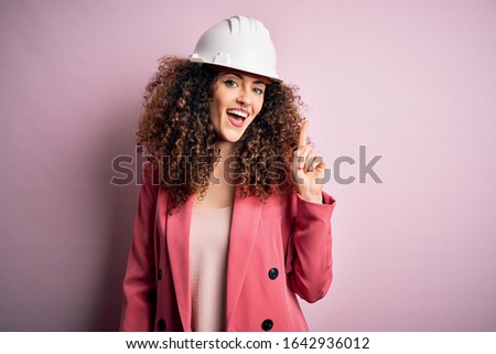Young beautiful architect woman with curly hair wearing safety helmet over pink background pointing finger up with successful idea. Exited and happy. Number one.