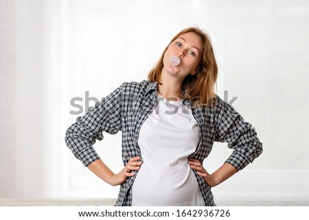 A young pregnant woman shows off her tummy and inflates a bubble of gum. White window in the background. Concept of pregnancy and motherhood. Close up