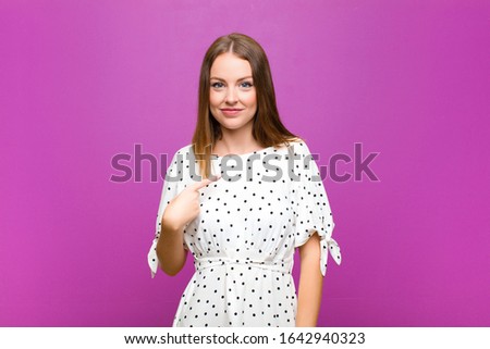 red head pretty woman looking proud, confident and happy, smiling and pointing to self or making number one sign against purple wall