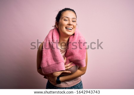 Young beautiful brunette sportswoman wearing sportswear and towel over pink background smiling and laughing hard out loud because funny crazy joke with hands on body.
