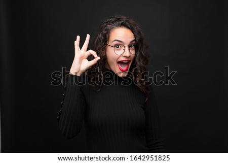 young pretty woman feeling successful and satisfied, smiling with mouth wide open, making okay sign with hand against black background.