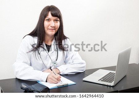 Doctor woman at work sitting at the desk in hospital or clinic, white coat, table with laptop and clipboard