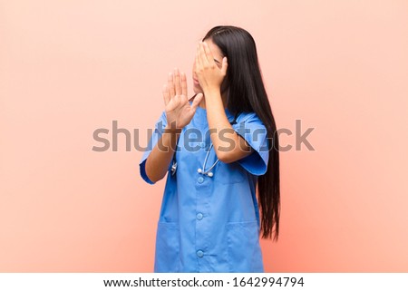 young latin nurse covering face with hand and putting other hand up front to stop camera, refusing photos or pictures against pink wall