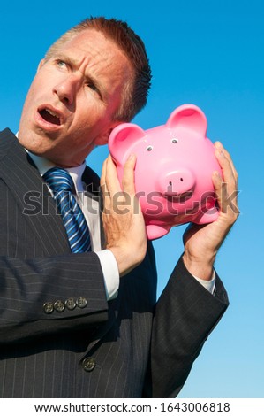 Frustrated businessman shaking a pink piggy bank listening for coins outdoors in bright blue sky