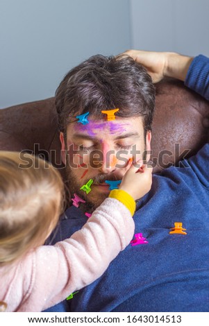 Young father suffering the mischief of his little daughter. Little girl paints with brush and watercolors her father's face while he sleeps on the couch Royalty-Free Stock Photo #1643014513
