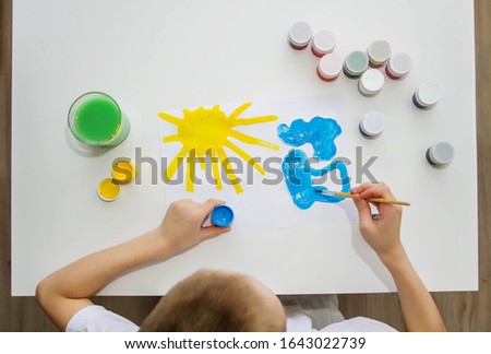 A boy in a white T-shirt at a white table paints the sun and clouds.