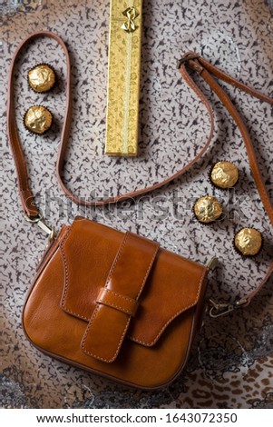 brown leather vintage bag and golden gift on a beige background with brown spots, a gift of gold and vintage leather bag top view, ladies leather bag and candy in gold foil and a gift of gold top view