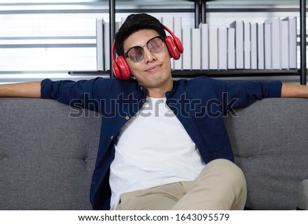 Relaxing Asian man sitting on sofa / couch listening to music / podcast via wireless headphone. Asian man wearing headphone / headset while relaxing in living room. Podcast  & happy lifestyle concept