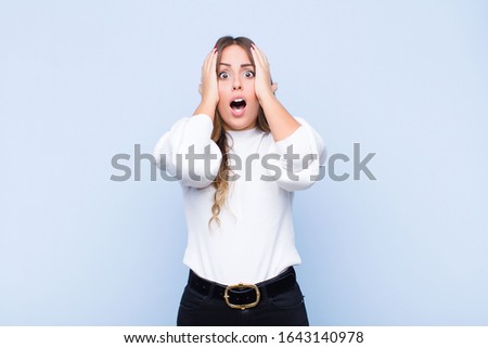 young pretty hispanic woman looking unpleasantly shocked, scared or worried, mouth wide open and covering both ears with hands against blue wall