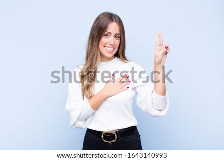 young pretty hispanic woman looking happy, confident and trustworthy, smiling and showing victory sign, with a positive attitude against blue wall