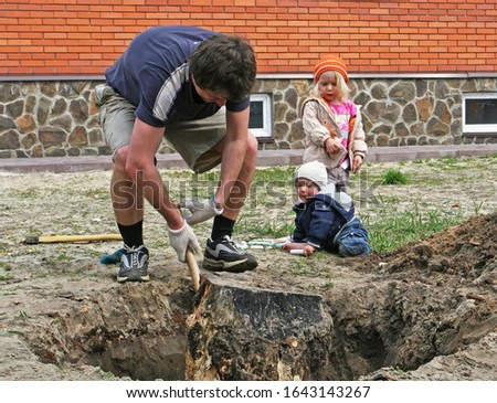 happy gardener dad uproots an old stump from the ground on a plot, farmer’s children are watching, spring has come, horizontal photo