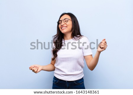young latin pretty woman smiling, feeling carefree, relaxed and happy, dancing and listening to music, having fun at a party against flat wall