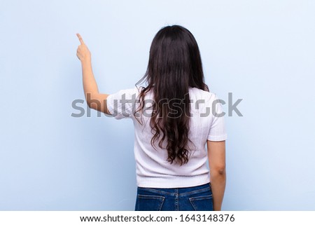young latin pretty woman standing and pointing to object on copy space, rear view against flat wall