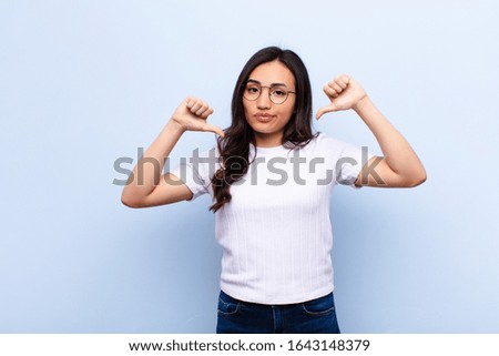 young latin pretty woman looking sad, disappointed or angry, showing thumbs down in disagreement, feeling frustrated against flat wall