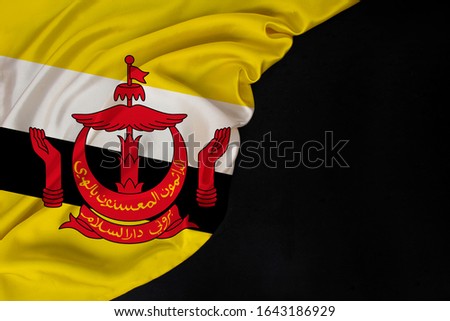 national flag of the state of Brunei on delicate shiny silk, inscription: Eternal service to Allah, folded onto black blank form, concept of tourism, economy, politics