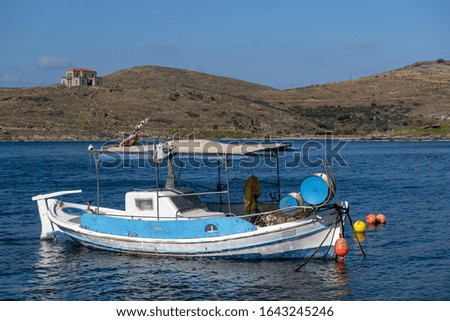 Tzia, Kea, Greece. Blue and white traditional fishing boat moored in the middle of calm sea at Vourkari cove. Mountains, buildings and blue sky background.