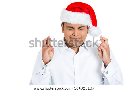 Closeup portrait of handsome young man wearing red santa claus hat crossing  fingers, closing eyes, wishing praying for a miracle, isolated on white background, space to left. Positive emotion
