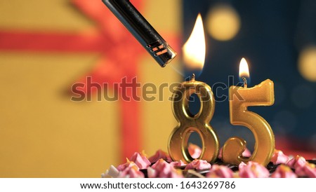 Birthday cake number 85 golden candles burning by lighter, background gift yellow box tied up with red ribbon. Close-up view Royalty-Free Stock Photo #1643269786