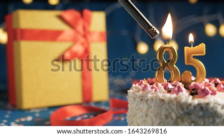 White birthday cake number 85 golden candles burning by lighter, blue background with lights and gift yellow box tied up with red ribbon. Close-up view Royalty-Free Stock Photo #1643269816