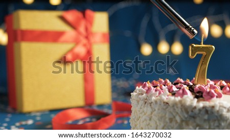 White birthday cake number 7 golden candles burning by lighter, blue background with lights and gift yellow box tied up with red ribbon. Close-up view