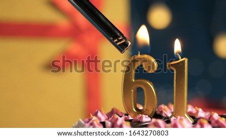 Birthday cake number 61 golden candles burning by lighter, background gift yellow box tied up with red ribbon. Close-up view