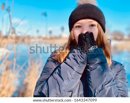 A surprised teenage girl in winter clothes  closing her face with the hands in gloves by the river