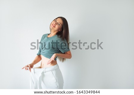 The joyful girl who lost weight points to her former huge pants. Woman Losing Weight After Diet, Slim Down, Burning Fat and Flat Waist. Thin Girl Show Weight Loss and Old Big Jeans. Too Large Pants.  Royalty-Free Stock Photo #1643349844