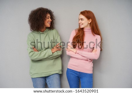 Studio photo of charming cheerful young ladies looking positively on each other and smiling happily, keeping hands crossed on chest while posing over grey background