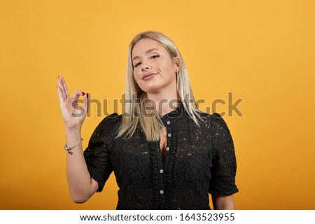 Young blonde girl in black dress on yellow background happy woman shows OK sign with her hands