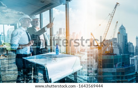 Two Engineer or Architect are analyzing blueprints while working on a new project on construction site with blue sky and city background.Architect supervising construction on terrace tower. Royalty-Free Stock Photo #1643569444