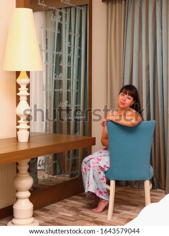 A 35-year-old brunette in a long dress sits on a blue chair in a hotel room near a table with a floor lamp, a large mirror with welcome signs Welcome in various languages