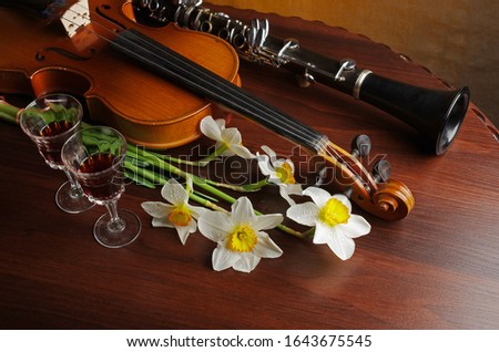Violin with a bow, a bouquet of white daffodils, clarinet and two glasses of wine on a wooden table.