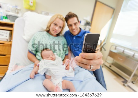 Man with woman and newborn babygirl taking selfportrait through cell phone in hospital room
