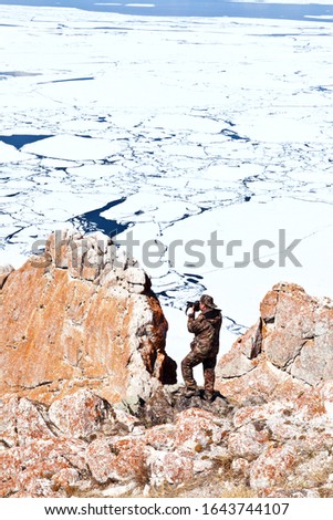 Baikal lake in spring. Red coastal cliffs of Olkhon Island against the background of ice drift in the Small Sea Strait. A tourist in a hiking suit photographs the landscape from Cape Sagan-Khushun 