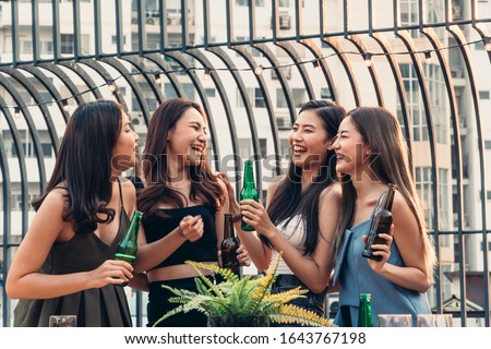 Group of happy Asian girl friends celebrating party with beer bottle toasting drinks at rooftop cafe in sunset together, female gang chatting, laughing on smile face, night lifestyle of young people